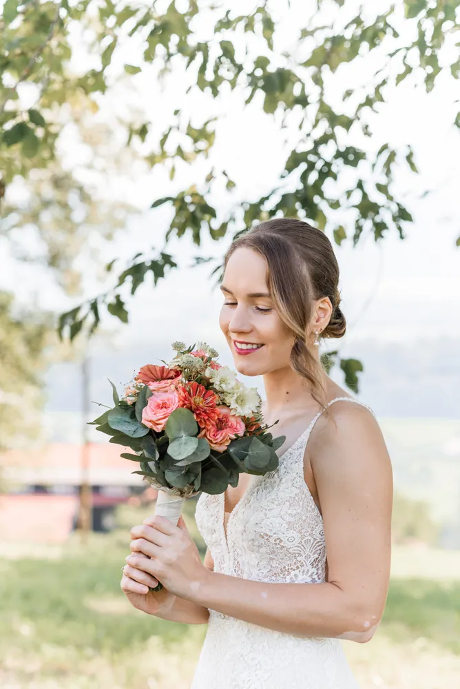 Bride in white lace dress holds bouquet of pink and white flowers, smiling outdoors with trees and building in background.