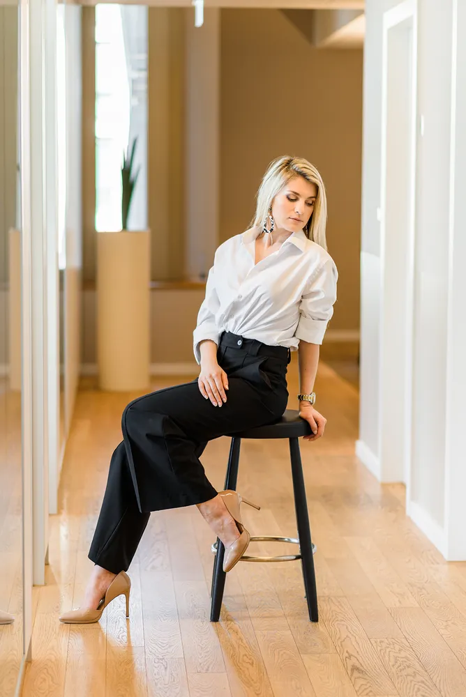 Woman in white shirt and black trousers seated on stool in modern hallway, high heels, contemplative expression.