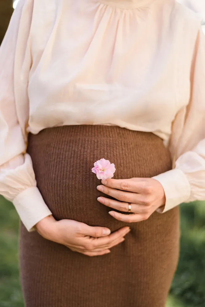 Pregnant woman in cream blouse and brown ribbed skirt, cradling belly with hands, holding pink flower.