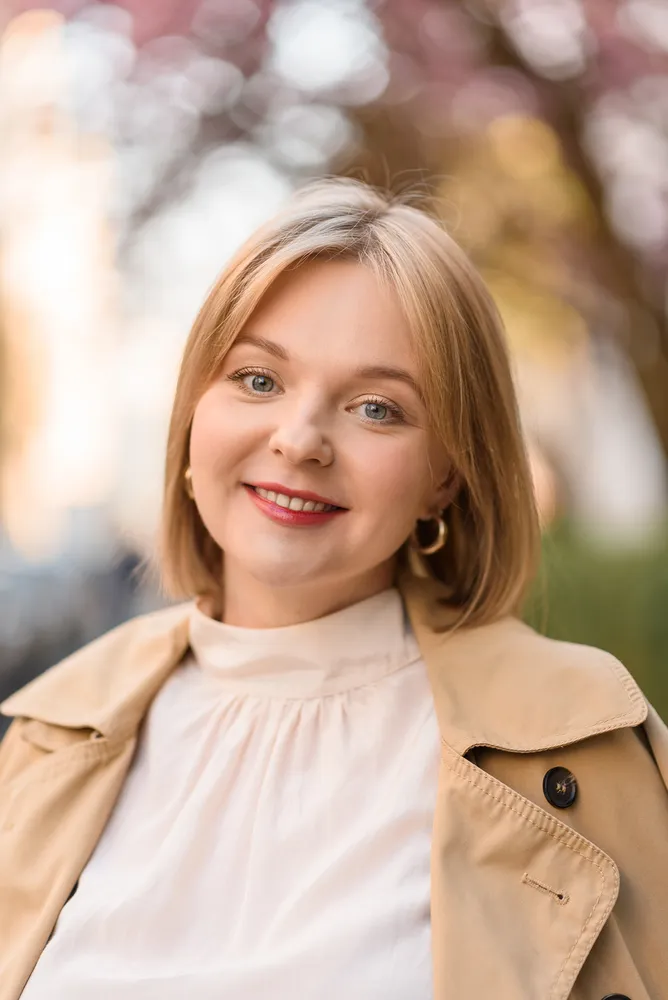 Woman in tan trench coat smiles outdoors. Blurred trees and sky in background.