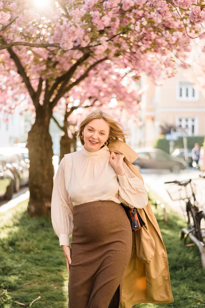 Woman strolls under blooming cherry tree, holding tan coat. She wears white blouse, brown skirt, smiles at camera. Urban street scene blurred in background.