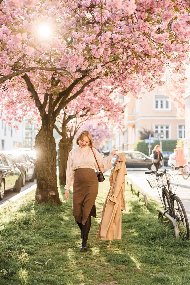 Woman in white blouse and brown skirt walks under pink cherry blossoms, holding tan coat, beside parked bicycle on grassy sidewalk.