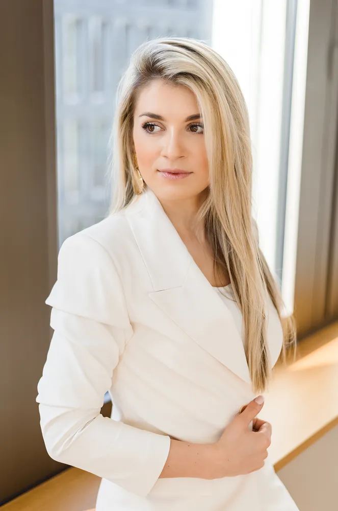 Woman in white blazer seated by window, hand on chest, long blonde hair, light makeup, professional setting.