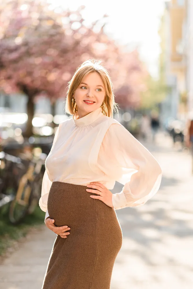 Pregnant woman in white blouse and brown skirt poses outdoors, hand on belly, spring blossoms in background.