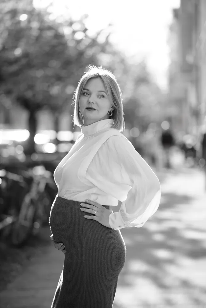 Pregnant woman stands confidently on a sunlit street, wearing a white blouse and dark skirt, her hand resting on her belly, gazing thoughtfully into the distance.