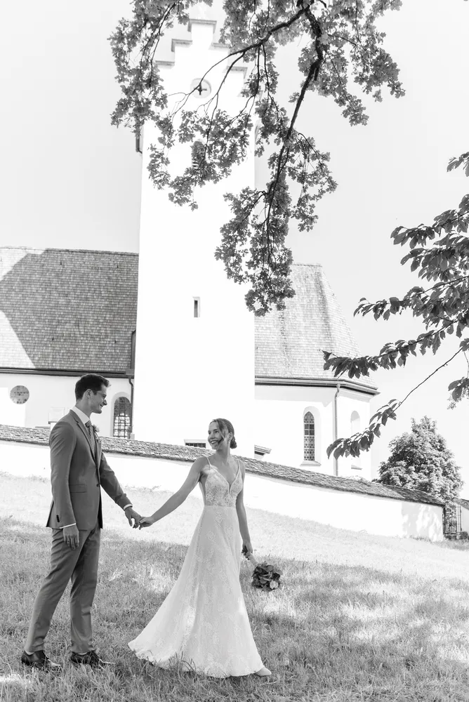 Couple in wedding attire holding hands, standing in a grassy field with a white church and trees in the background, black and white photograph.