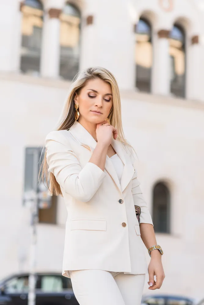 Woman in white suit adjusts blazer collar outdoors, gold watch and earrings visible, blurred urban background.