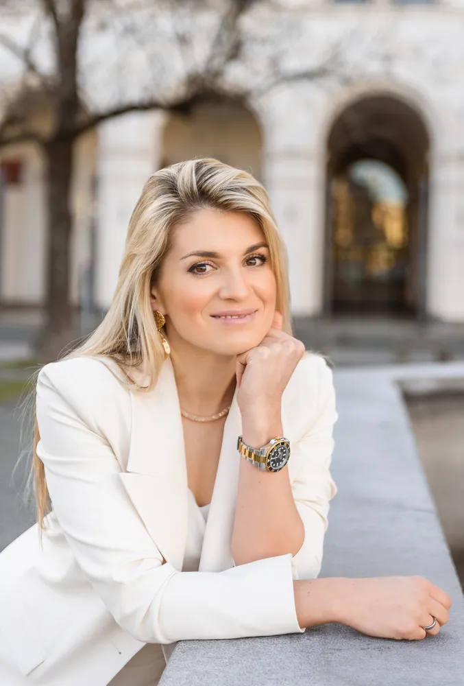 Woman in white blazer leans on ledge, gazes forward. Gold jewelry, watch, and ring adorn her. Outdoor, blurred building in background.