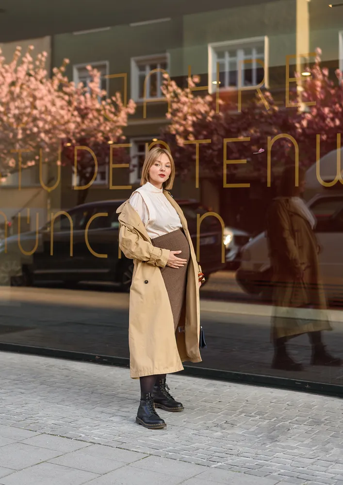 Pregnant woman in tan trench coat, white blouse, brown skirt, and black boots stands on cobblestone sidewalk, hand on belly, facing camera, reflection visible in storefront window behind her.