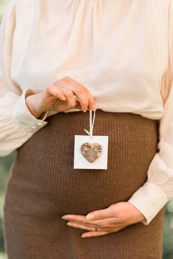 Pregnant woman in beige blouse and brown skirt holds wooden heart ornament with string, outdoors.