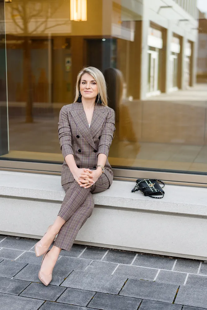 Woman in plaid suit seated on bench outside building, handbag beside her.