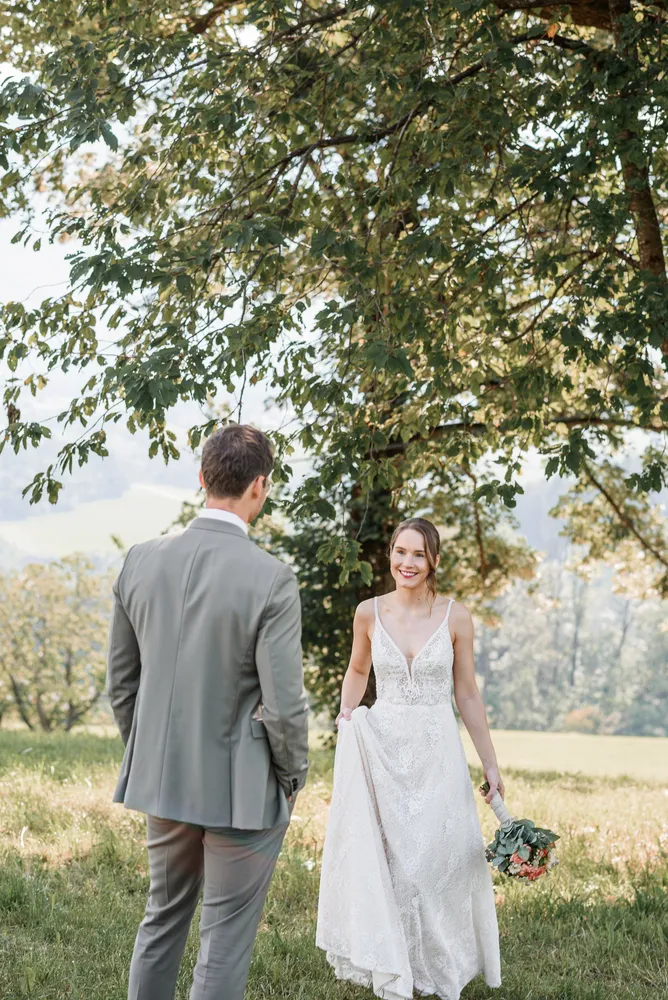Bride in lace wedding dress smiles while holding bouquet, groom in gray suit faces her, under large tree with expansive green landscape in background.