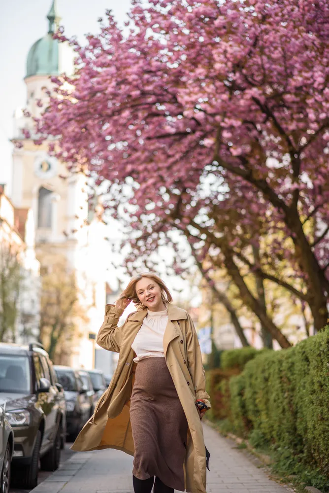 Woman in beige trench coat walks on sidewalk, smiling, hand on head. Pink blossoms overhead. Parked cars line street. Blurred historic building in background.
