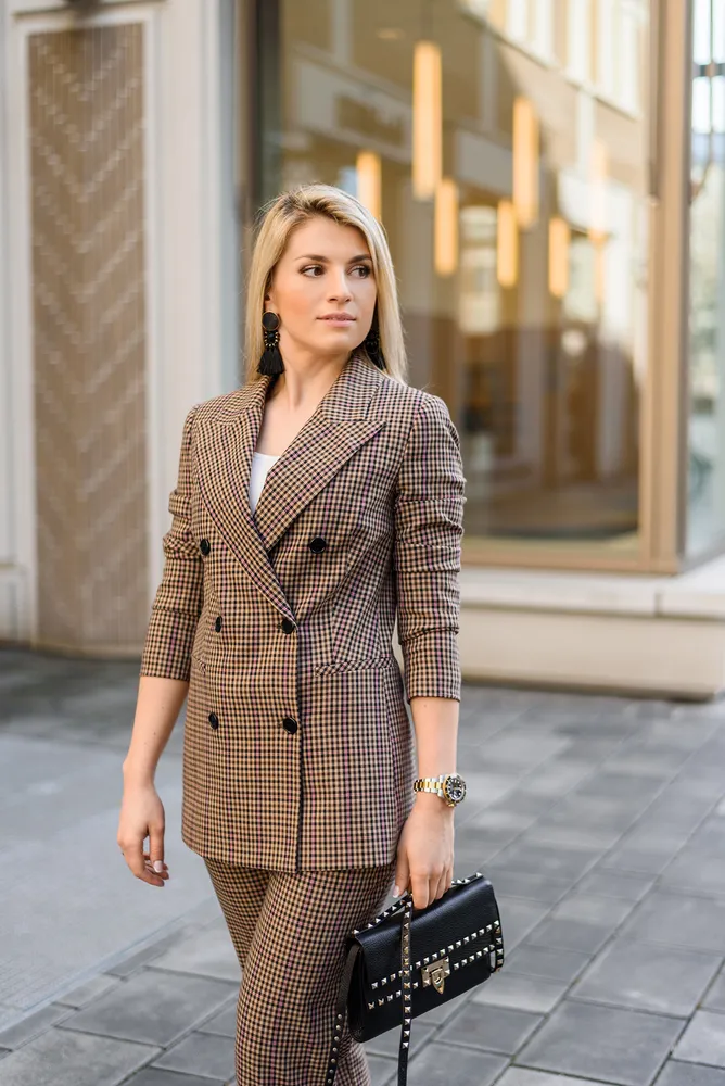 Woman in brown plaid suit and white top, wearing large earrings and watch, carries black studded purse, standing on stone pavement outside building with glass facade.