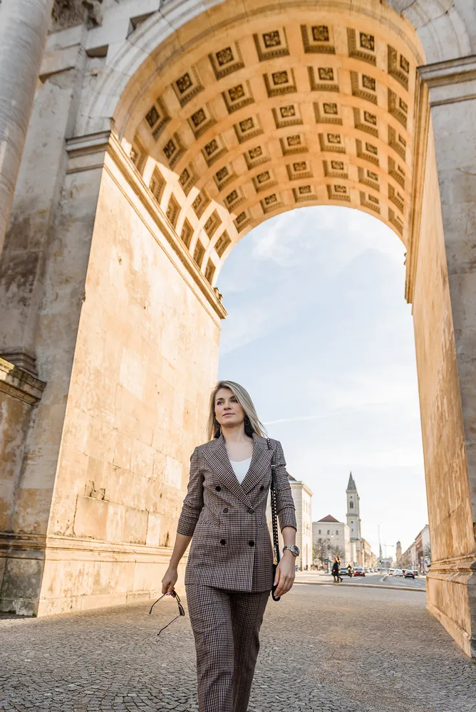 Woman in business attire walks through historic stone archway, holding sunglasses, with cobblestone street and distant buildings visible.