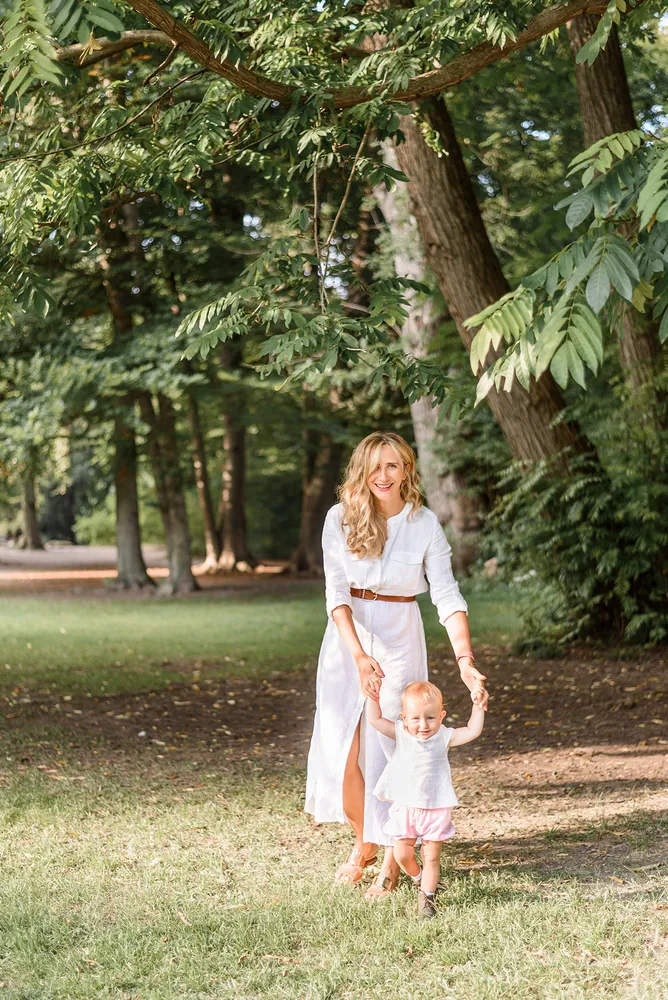 Woman in white dress walks with toddler holding hands in sunlit park. Green trees frame scene. Both smiling. Grass underfoot.