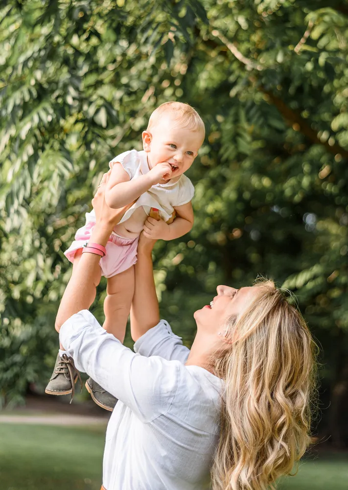 Woman in white blouse joyfully lifts toddler in pink shorts and white shirt against blurred green foliage backdrop.