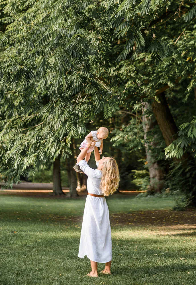 Woman in white dress joyfully tosses toddler in air amidst lush park greenery.
