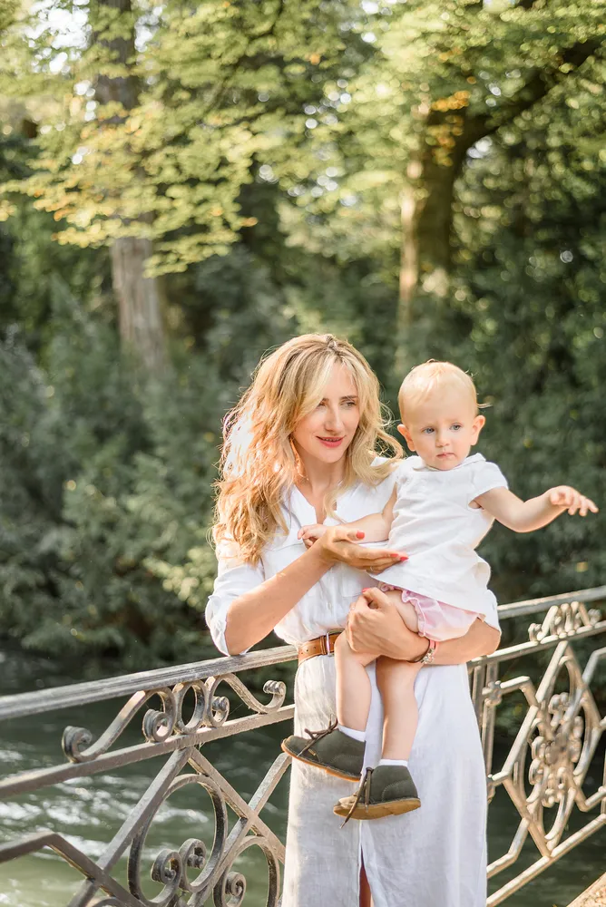 Woman in white dress holds child on ornate bridge overlooking calm water, surrounded by lush greenery.