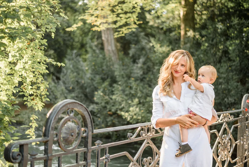 Woman and child in white dresses on a bridge, surrounded by greenery. Sunlit, candid moment captured.