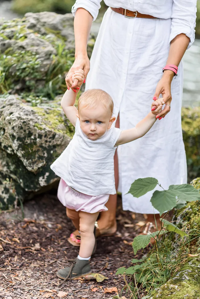 A woman in a white dress and a young child in a white shirt and pink skirt hold hands on a gravel path surrounded by greenery and rocks.