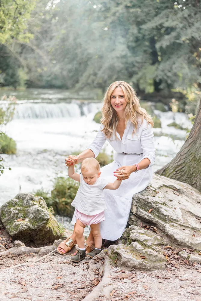 Woman in white dress holds toddler's hands by a waterfall. Both smiling. Natural outdoor setting with trees and rocks. Soft, diffused lighting.