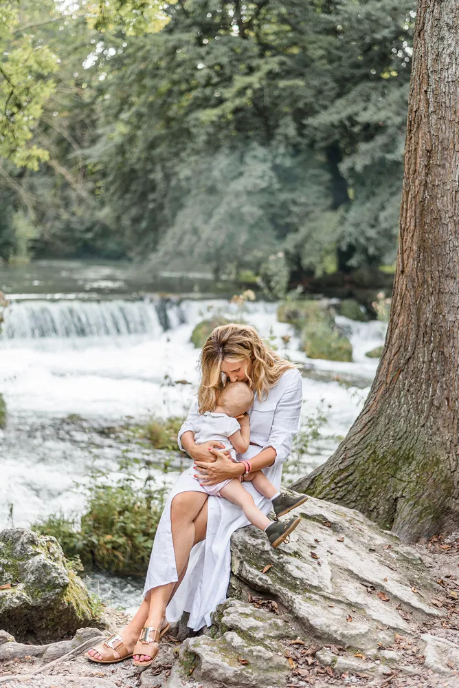Woman in white dress cradles baby against tree beside flowing stream, serene forest backdrop.