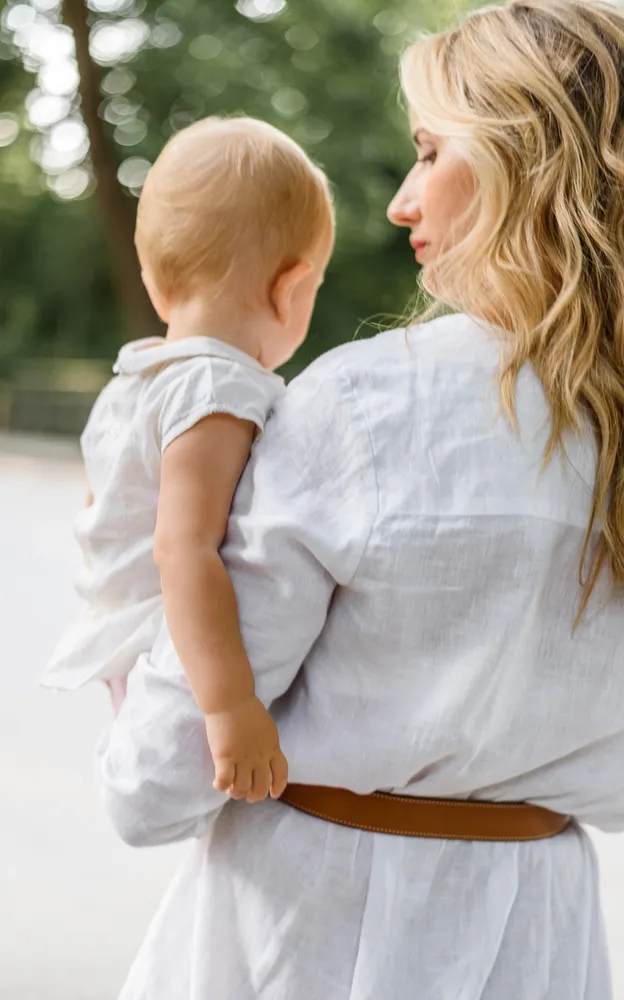 Woman with blonde hair in white blouse cradles a small child in white outfit. They stand outdoors, surrounded by greenery, facing away from camera. Soft focus background.