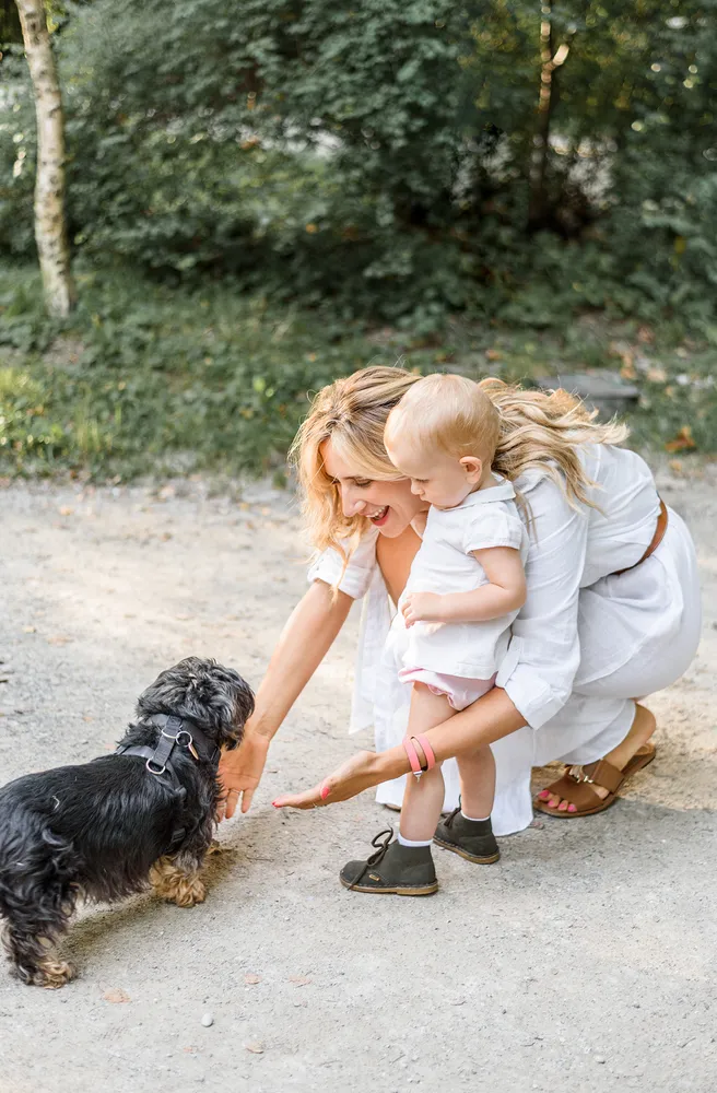 Woman in white dress squats on gravel, extending hand to small black dog wearing harness. Child in pink shorts clings to her back. Both have pink-painted nails. Blurred greenery in background.