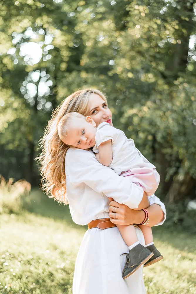 A woman with long blonde hair and a child embrace in a sunlit park, both dressed in white, surrounded by blurred greenery.