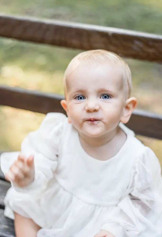 A young toddler with light hair and blue eyes, wearing a white dress with lace sleeves, sits on a dark wooden bench. The child is waving one hand while looking directly at the camera, set against a blurred outdoor background.