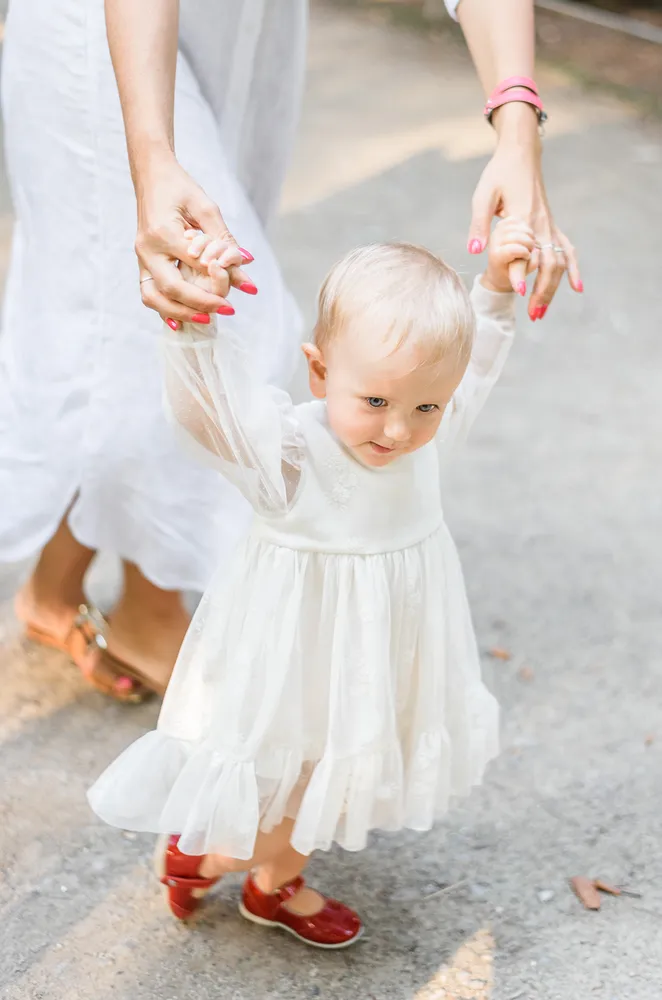 A toddler in a white lace dress and red shoes walks, holding an adult's hands. The adult wears white pants and pink accessories. They walk on a gravel path, outdoors.