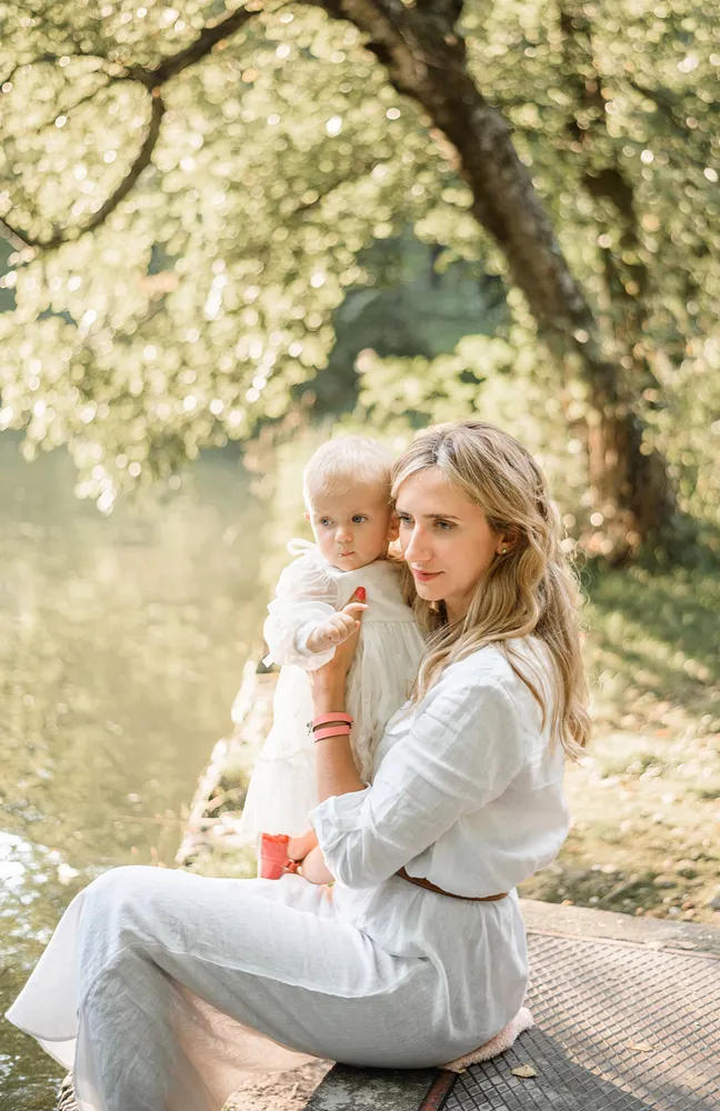 Woman in white dress sits by a tranquil lake, cradling a toddler in a white dress. Both gaze softly towards the camera, surrounded by a sunlit, leafy woodland backdrop. The serene setting suggests a peaceful, intimate moment captured in nature.