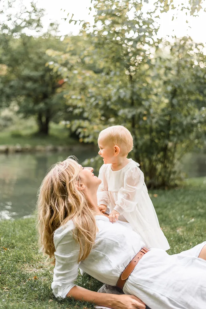 Mother and child in white dresses share a joyful moment by a serene lake, surrounded by lush greenery. The mother lies on the grass, gazing up at her child who stands beside her, both smiling.