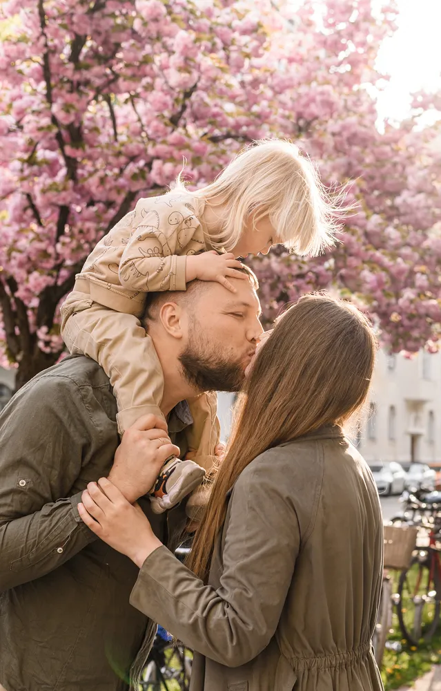 A family shares a kiss under a blooming tree, man carrying child on shoulders.
