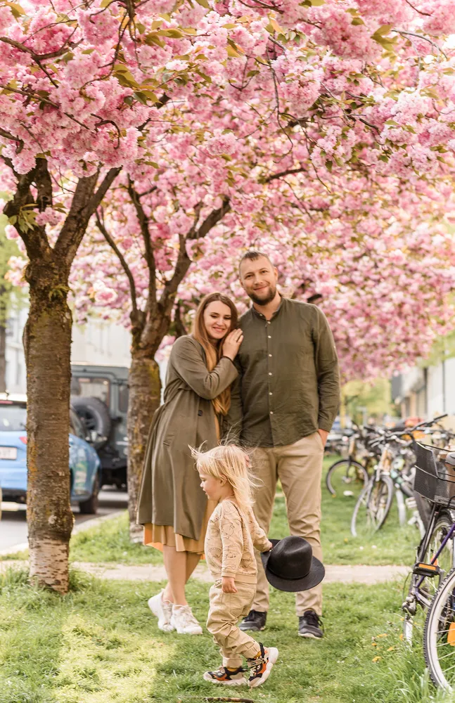 A family of three smiles under a canopy of pink blossoms. The mother embraces the father while their young daughter, holding a hat, walks towards them. Parked cars and bicycles line the background, framing the joyful scene.