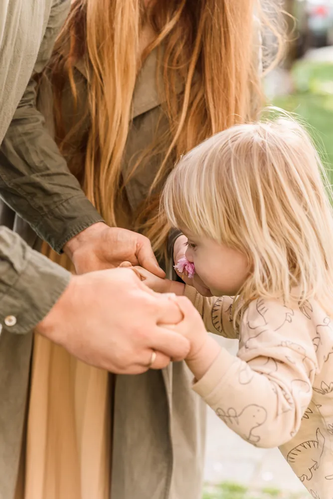 A woman with long auburn hair, wearing a beige dress, is seen bending down to a child with blonde hair. The child, dressed in a light-colored onesie with a dinosaur pattern, is holding a pink flower close to their nose. The woman’s hand is gently resting on the child’s hand, guiding their attention to the flower. The background is blurred, but hints of greenery suggest an outdoor setting. The image captures a moment of shared curiosity and gentle interaction between the two subjects
