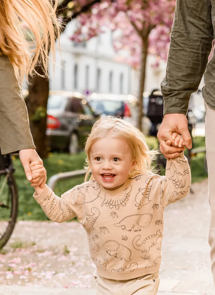 A young child with blonde hair wearing a beige dinosaur-patterned shirt walks between two adults on a tree-lined street with parked cars and cherry blossoms in the background.