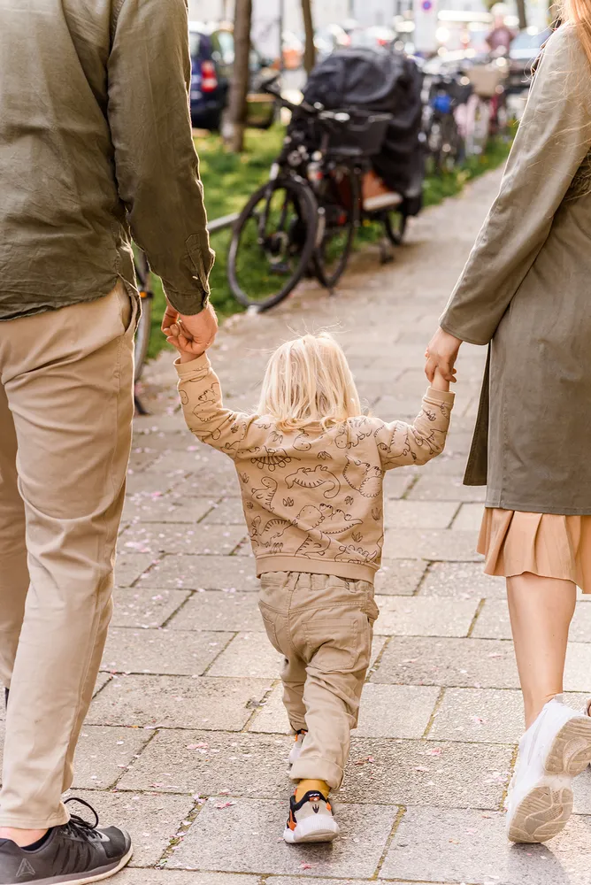 A toddler walks down a sidewalk, holding hands with two adults, surrounded by parked bicycles and trees.
