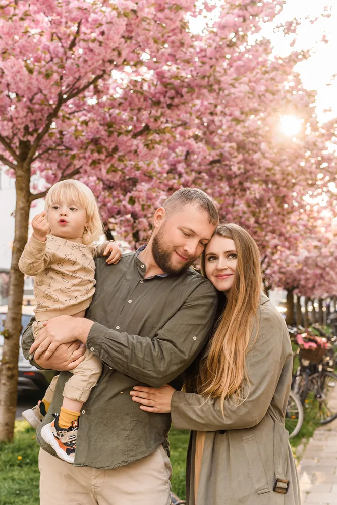 A family of three smiles outdoors, embracing under blooming cherry trees. The father holds a child, while the mother leans in, all dressed in casual attire.