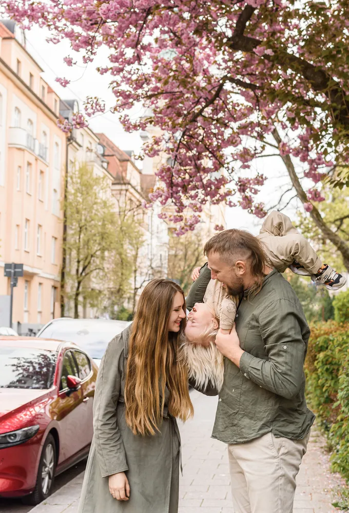 Family portrait: A man with long hair joyfully holds a child above his head as a woman with long hair stands beside them, smiling. They are on a sidewalk with buildings and cherry blossoms in the background.