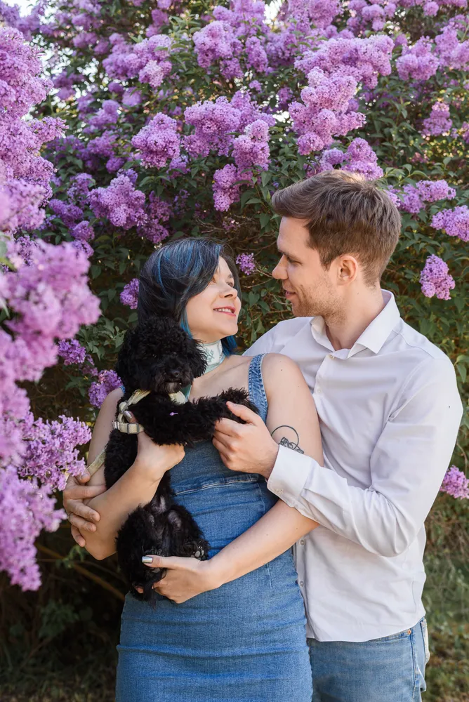 Couple embracing outdoors, woman holding small black dog, surrounded by purple lilacs, both smiling, casual attire.