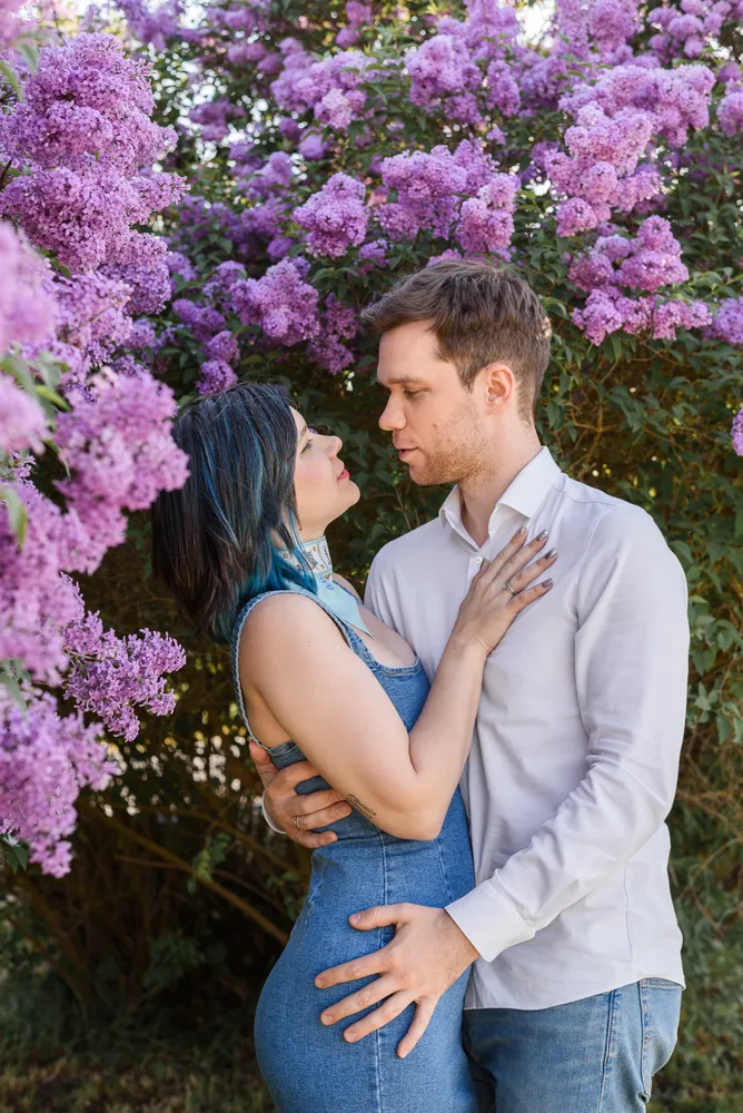 A couple embraces amidst blooming lilac bushes. The woman, with dyed hair, wears a blue dress; the man, in a white shirt and jeans, gazes at her. Their hands gently touch. Sunlit petals frame their affectionate pose.