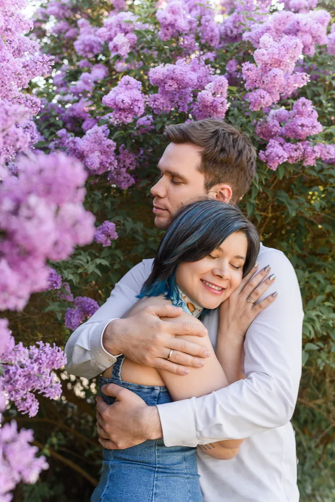 Couple embracing in a park surrounded by blooming lilac bushes. Woman with blue-tipped hair and man in white shirt. Romantic, intimate moment captured.
