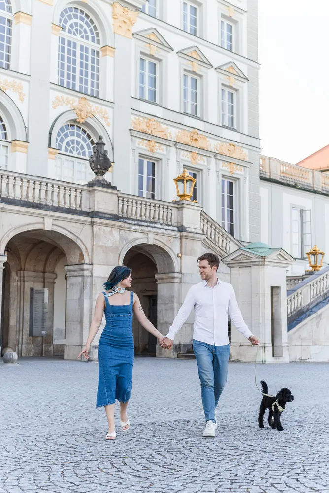 A man and woman in casual attire hold hands while walking a small black dog on cobblestone pavement in front of an ornate historic building with arched doorways and gold accents.
