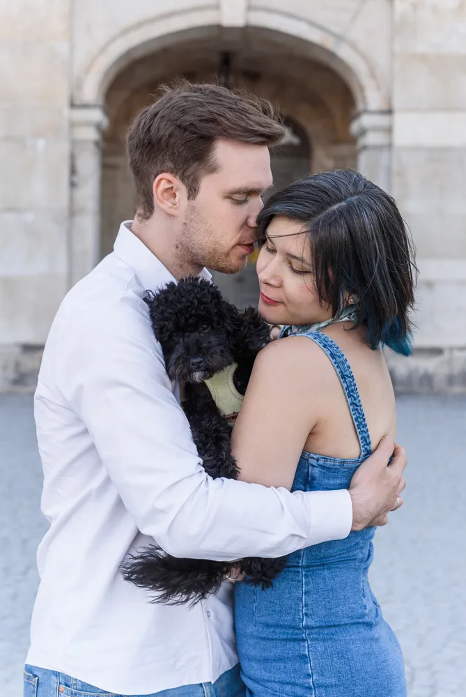 Couple embracing with small black dog, urban backdrop, affectionate moment captured.