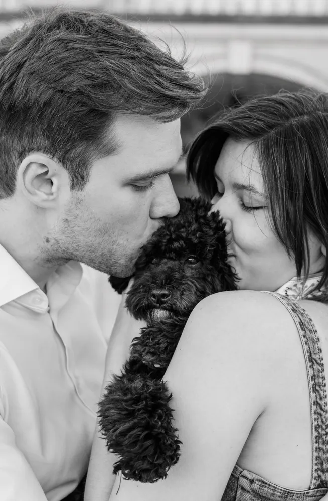 A man and woman share a tender moment with a small black poodle in a black and white photograph, their faces close together as they kiss the dog's head. They are dressed casually, with the man in a light-colored shirt and the woman in a sleeveless top, set against an indistinct outdoor backdrop.