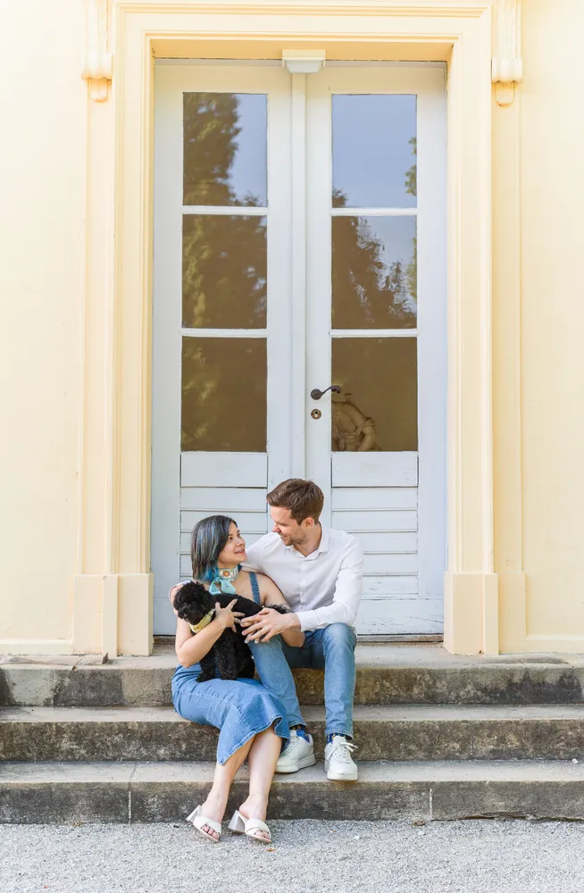 Couple with small dog sits on stone steps in front of white double doors with glass panels, smiling and embracing. Woman wears blue dress, man in white shirt and jeans. Sunlit exterior, neutral-toned building.