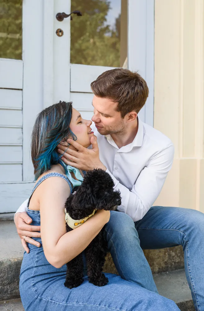 Couple sharing a kiss with a black poodle on their lap, seated on steps in front of a white door with glass panes, woman has blue-tipped hair, man in white shirt, both in blue jeans.