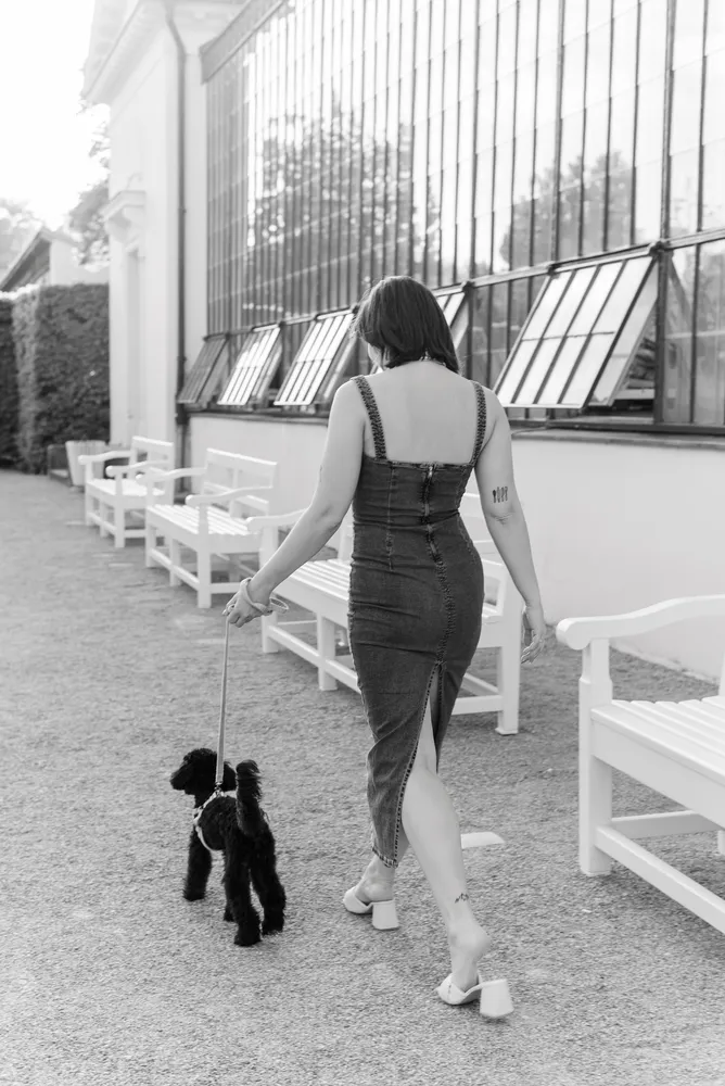 Woman in sleeveless dress walks a black poodle on a leash beside outdoor benches and a building with tall windows.